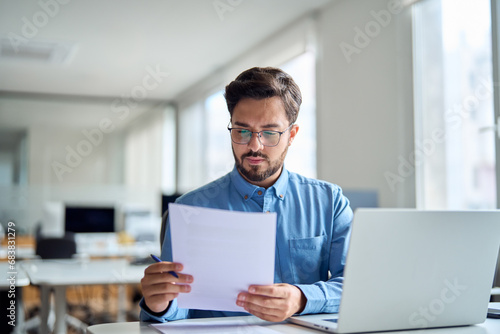 Young busy latin professional business man checking document working at laptop computer in office. Serious businessman accountant expert reading legal paper company file overview at workplace.