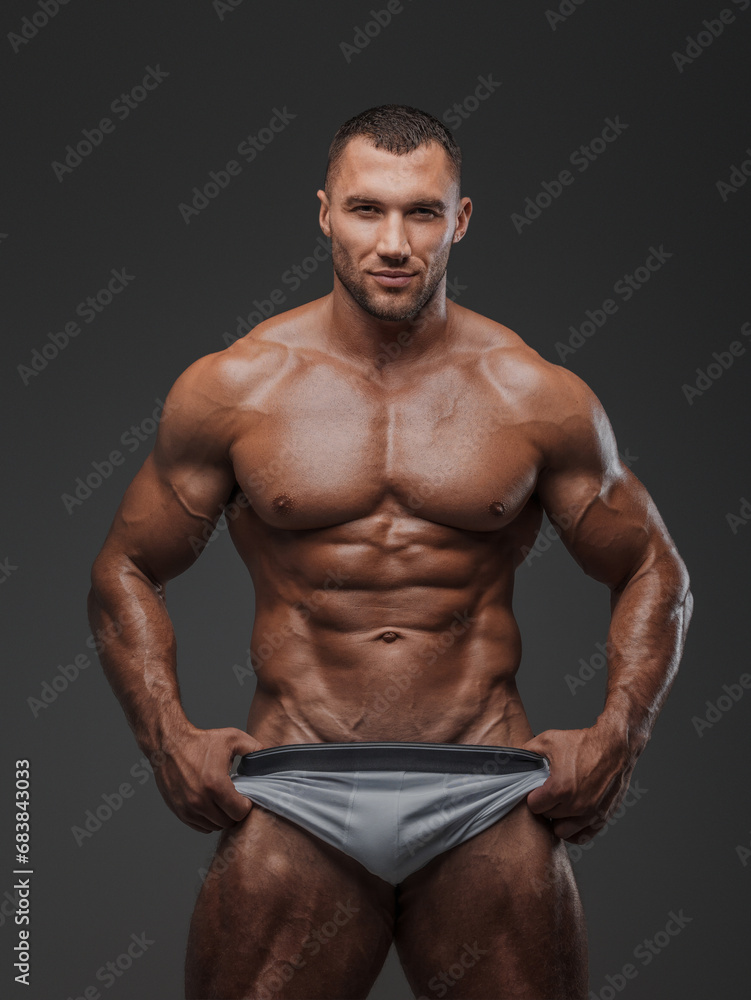 Portrait of a rugged man with a well-groomed model appearance, wearing white briefs, and showcasing his bare muscular torso against a gray background