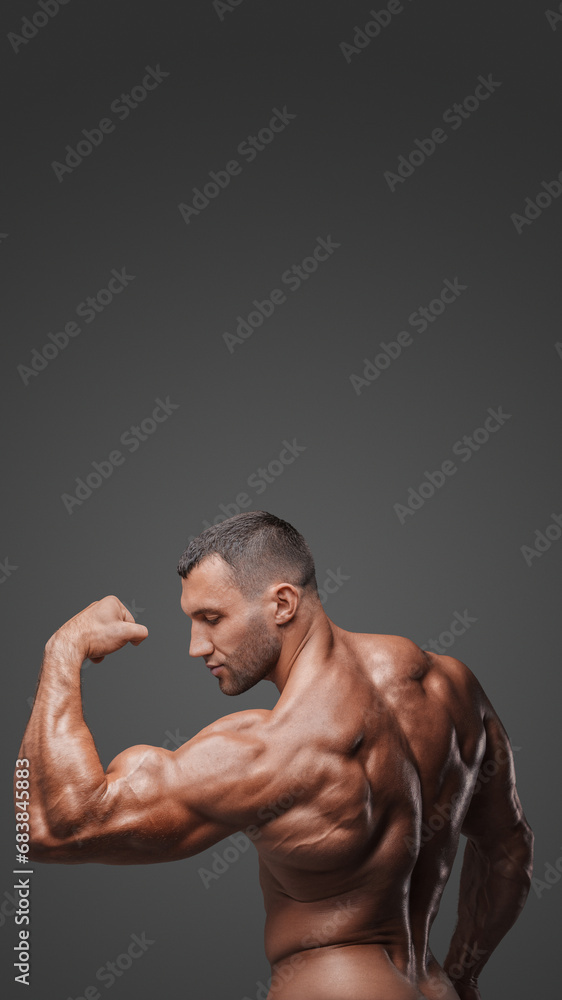 Fototapeta premium View from behind of a muscular man with a bare torso, striking a pose, showcasing his tense muscles against a gray background