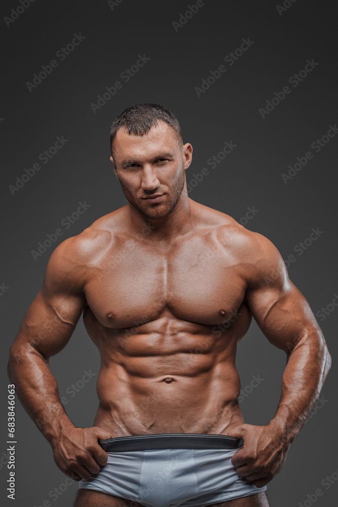 Portrait of a rugged man with a well-groomed model appearance, wearing white briefs, and showcasing his bare muscular torso against a gray background