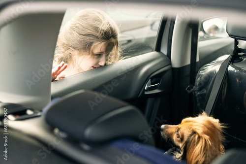 Little girl is looking through a window car to a cute little dog which is sitting inside the car © Florincristian