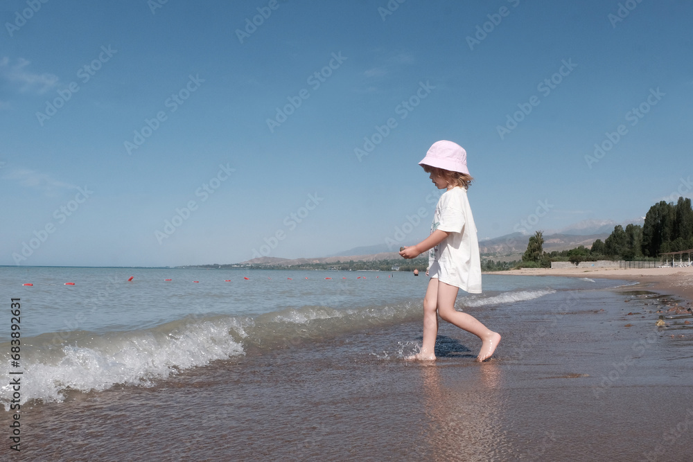 A little fair-haired girl in a Panama hat and white clothes is running along the seashore. Concept of safe water games, healthy lifestyle.