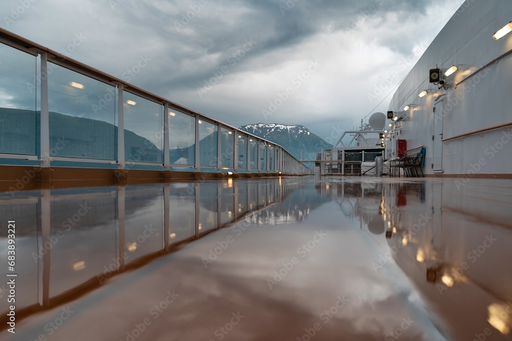 Reflection of Norwegian landscape in a puddle on the open deck on cruise ship, with fjords with snow, with clouds, with lights and handrails, with a bench, Lisafjord, Norway