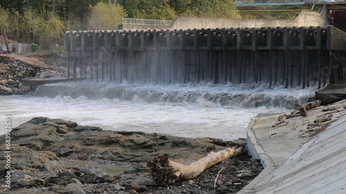 a dam with a powerful flow of water, a view from the shore with branches and trees applied, the power of the element of water in the design of a city dam with splashes and the pressure of a stream