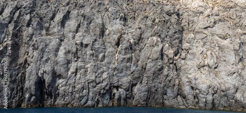 Weathered seaside rock face texture with parts of green and blue water. Aged volcanic stone wall surface background pattern with cracks and scratches. Banner. Ischia Island, Italy.