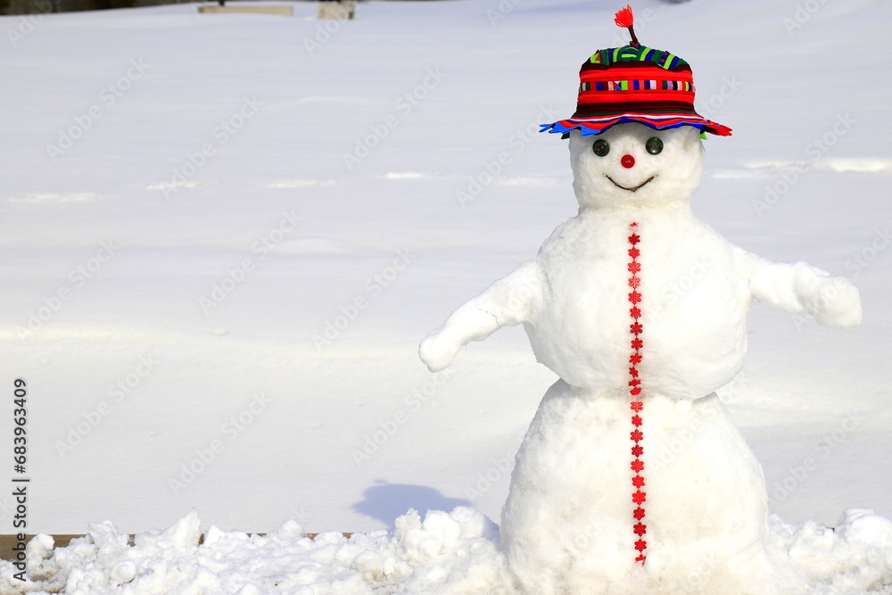 Obraz premium A funny snowman in red hat stands on snow covered meadow in winter, Christmas, New Year holidays, children entertainment and sports