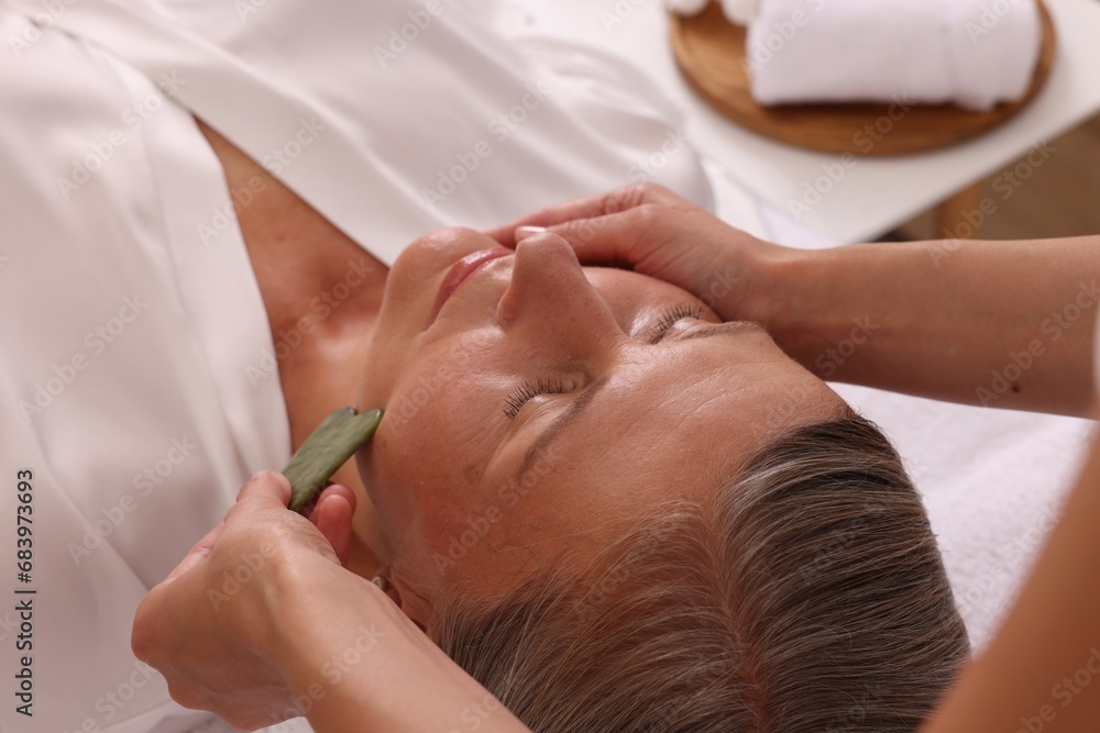 Woman receiving facial massage with jade gua sha tool in beauty salon, closeup Woman receiving facial massage with jade gua sha tool in beauty salon, closeup