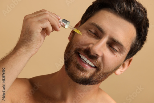 Smiling man applying cosmetic serum onto his face on beige background, closeup