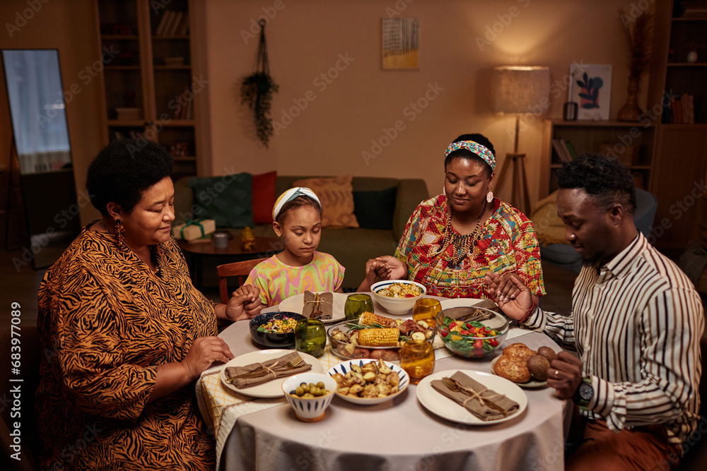 Black family sitting at Kwanzaa dinner table with eyes closed, hold hands and praying