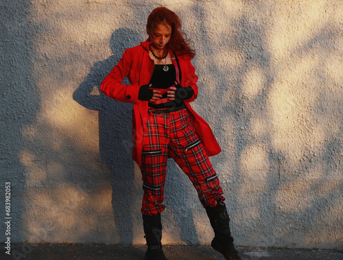 Selfie photo of a woman near a white gray wall in tight black clothes, a red raincoat, a leather bag. Cinematic punk style, fashion.