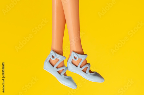 Slim and slender legs of a doll wearing light sandals on a yellow background.