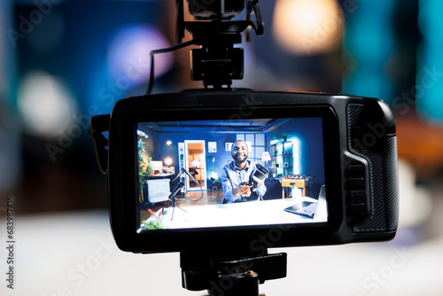 Focus on professional video production camera used for capturing footage of tech engineer in blurry background filming virtual reality product review for online audience, showcasing VR headset