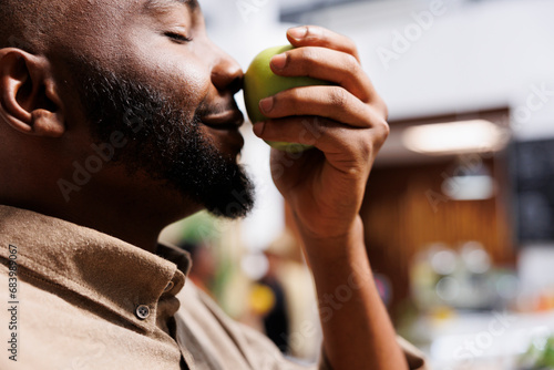 Selective focus on black man at zero waste store smelling an organic farm-grown apple. Close-up of African American male customer trying to determine if local neighborhood shop fruits are fresh.