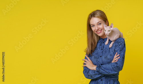 Smiling blond woman showcases unique bond with Egyptian cat Sphinx as it sits on shoulder against yellow background. Special companionship between woman and exotic feline friend, pet owner and cat. photo