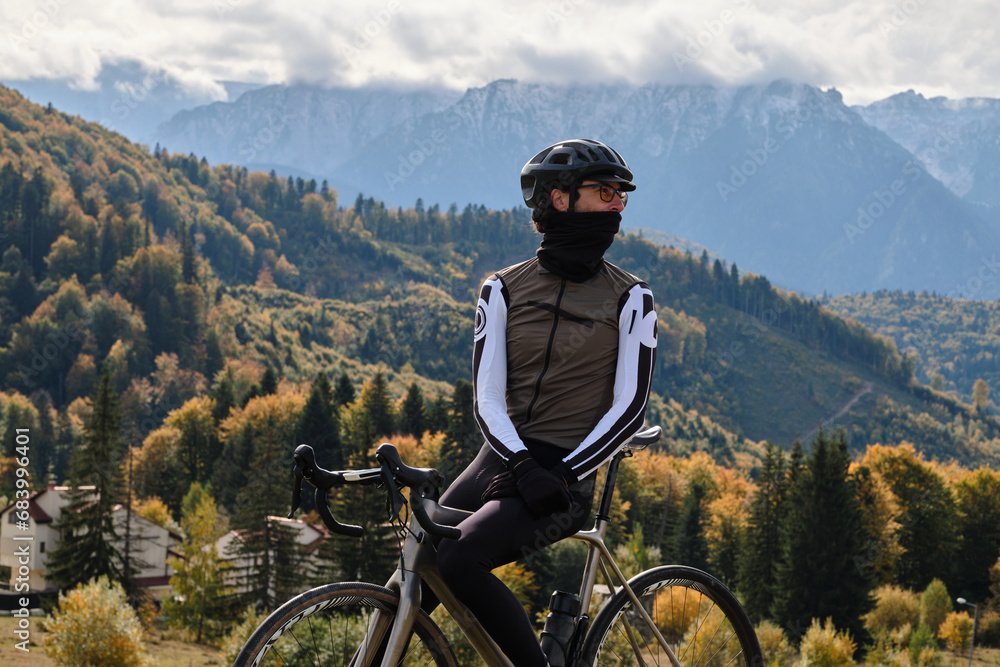 Fototapeta premium Close-up photo of man cyclist wearing full cycling winter kit and helmet. Man cyclist standing against autumn mountains background. Cyclist riding gravel bike. Gravel biking adventure in mountains.