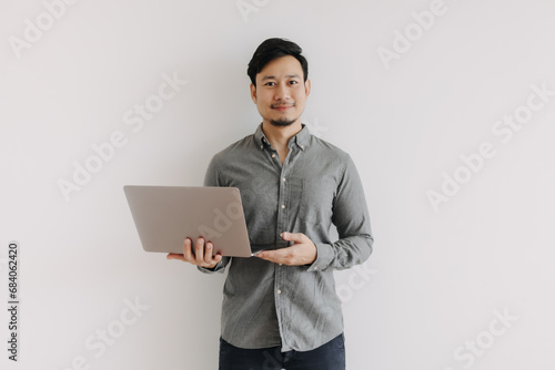 Asian man with beard wear grey shirt using and holding laptop, presenting notebook with happy smiling face, looking at camera and standing isolated over white background wall.