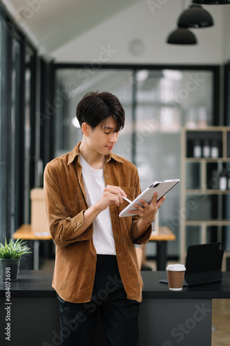 Confident Asian man with a smile standing holding notepad and tablet at office.