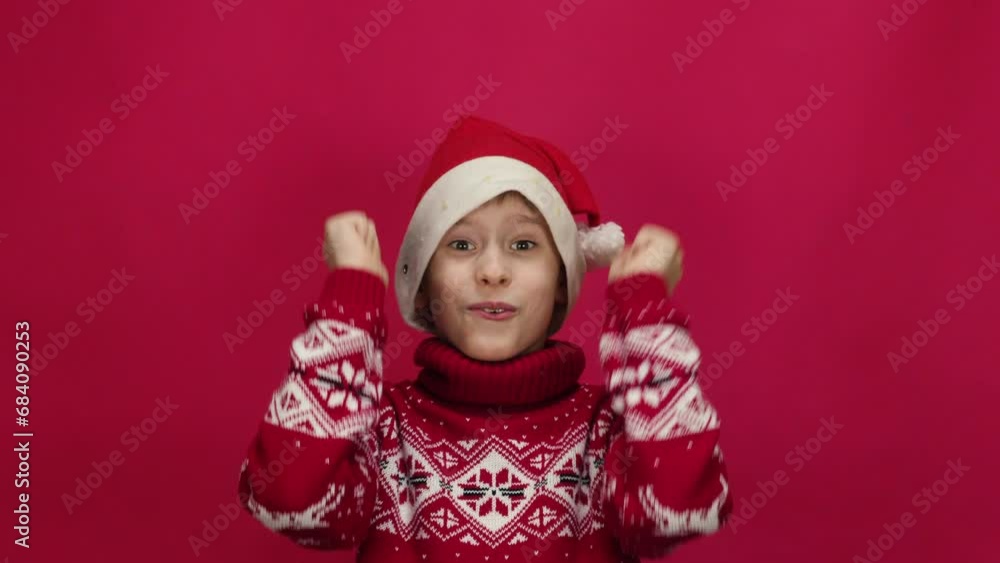 a joyful Caucasian schoolboy in a sweater and a Santa hat looks at the camera and shows a gesture of experience and success. studio shooting. red background.waiting for gifts and miracles a joyful Caucasian schoolboy in a sweater and a Santa hat looks at the camera and shows a gesture of experience and success. studio shooting. red background.waiting for gifts and miracles