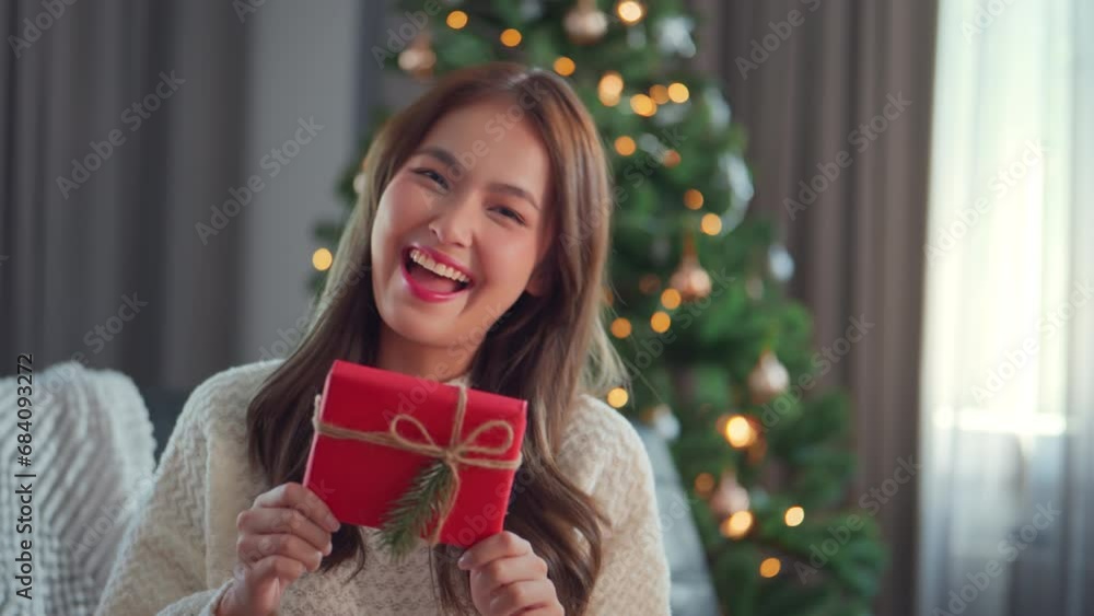 Happy attractive asian woman holding and showing a red gift to the camera. smile woman sitting near a decorated tree with Christmas decorations, Christmas Eve, New Year