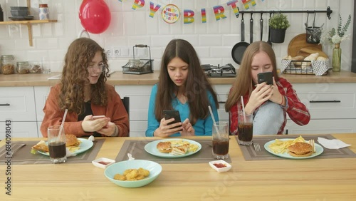 relaxed cheerful cute teenage female friends sitting at festive table, networking online on cellphones , talking and discussing social media content while celebrating birthday party indoors.