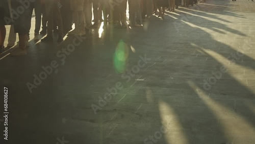 Shadows on ground cast by very long queue of unrecognizable people standing on street or city square at sunset, anamorphic shot.