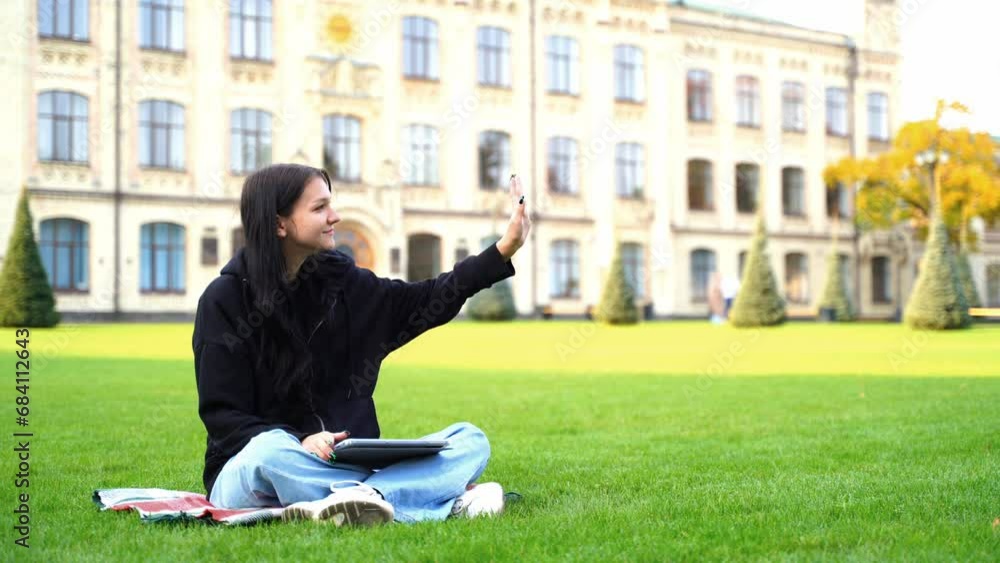 Portrait of a student girl studying and working online using a laptop on the lawn near the university. A young woman unexpectedly met an acquaintance and waves her hand smiling Portrait of a student girl studying and working online using a laptop on the lawn near the university. A young woman unexpectedly met an acquaintance and waves her hand smiling
