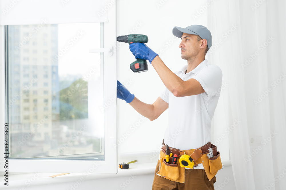 Using drill by installing windows. Repairman is working indoors in the modern room Using drill by installing windows. Repairman is working indoors in the modern room