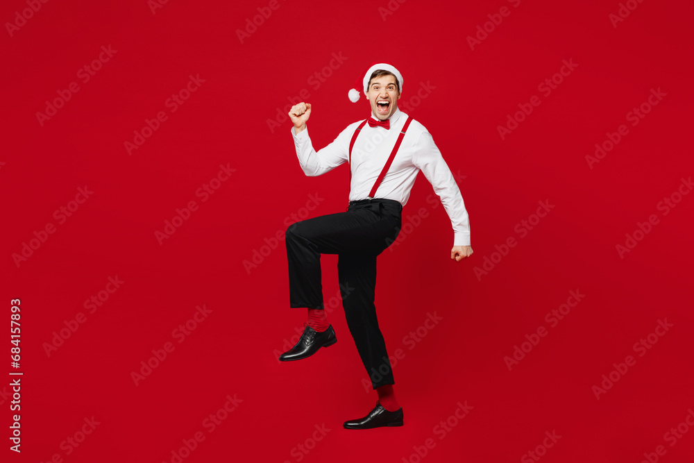 Full body side profile view merry young man wearing white shirt Santa hat posing do winner gesture raise up leg isolated on plain red background. Happy New Year Christmas celebration holiday concept. Full body side profile view merry young man wearing white shirt Santa hat posing do winner gesture raise up leg isolated on plain red background. Happy New Year Christmas celebration holiday concept.