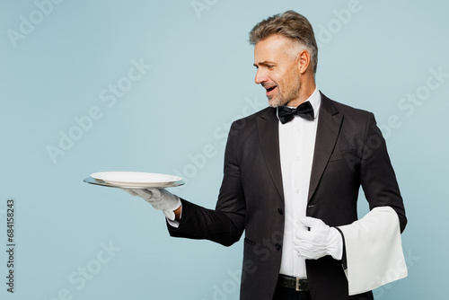 Adult sad barista male waiter butler man wear shirt black suit bow tie elegant uniform hold empty plate look camera towel work at cafe isolated on plain blue background. Restaurant employee concept. photo