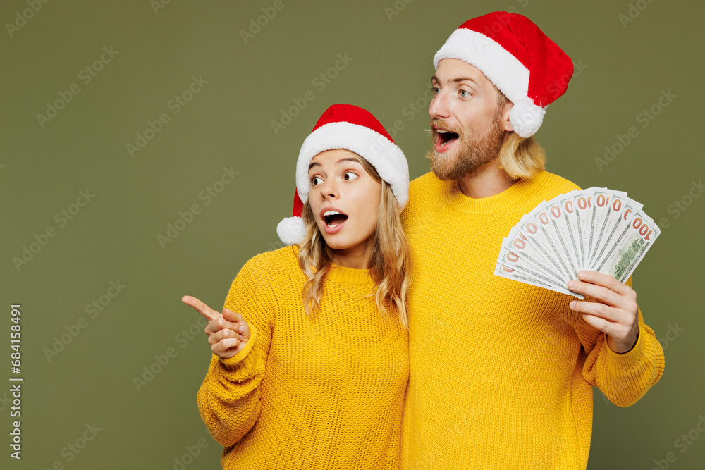 Merry young couple two friends man woman wear sweater Santa hat posing hold fan cash money in dollar banknotes point aside isolated on plain green background. Happy New Year Christmas holiday concept. Merry young couple two friends man woman wear sweater Santa hat posing hold fan cash money in dollar banknotes point aside isolated on plain green background. Happy New Year Christmas holiday concept.