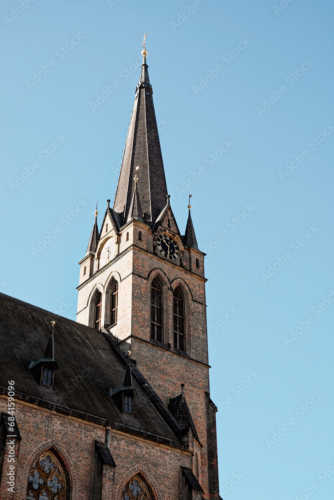 Obraz premium The image shows a brick church with a tall steeple and a clock on the tower. The church has arched windows and is lit by the sun against a blue sky. Spire on the bell tower of a Christian church.