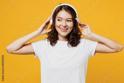 Young fun cheerful smiling happy woman wear white blank t-shirt casual clothes listen to music in headphones look aside isolated on plain yellow orange background studio portrait. Lifestyle concept.