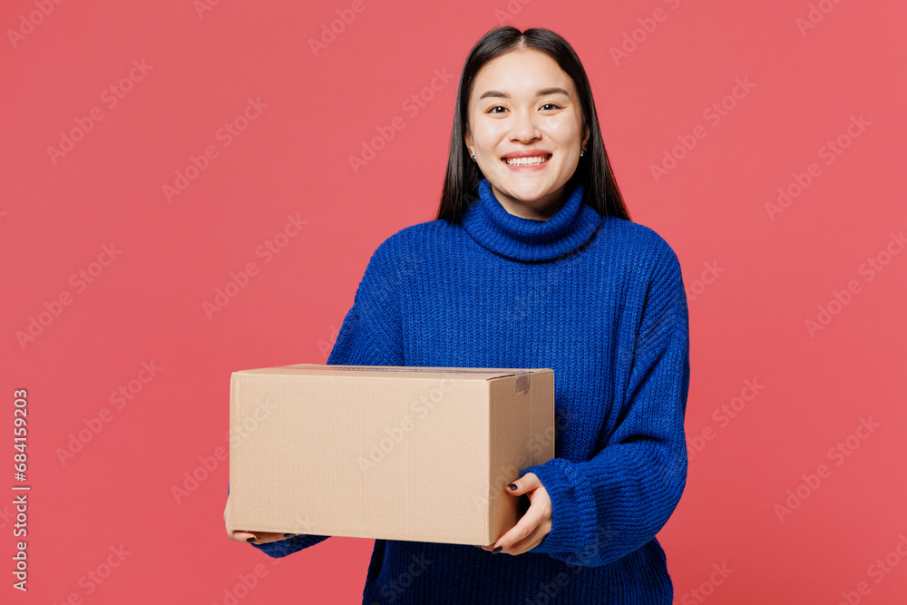 Young smiling happy woman of Asian ethnicity she wearing blue sweater casual clothes hold cardboard box look camera isolated on plain pastel light pink background studio portrait. Lifestyle concept. Young smiling happy woman of Asian ethnicity she wearing blue sweater casual clothes hold cardboard box look camera isolated on plain pastel light pink background studio portrait. Lifestyle concept.