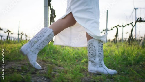 At sunset in the vineyard side walking in front of the camera lady wearing white cowboy boots and white dress photo