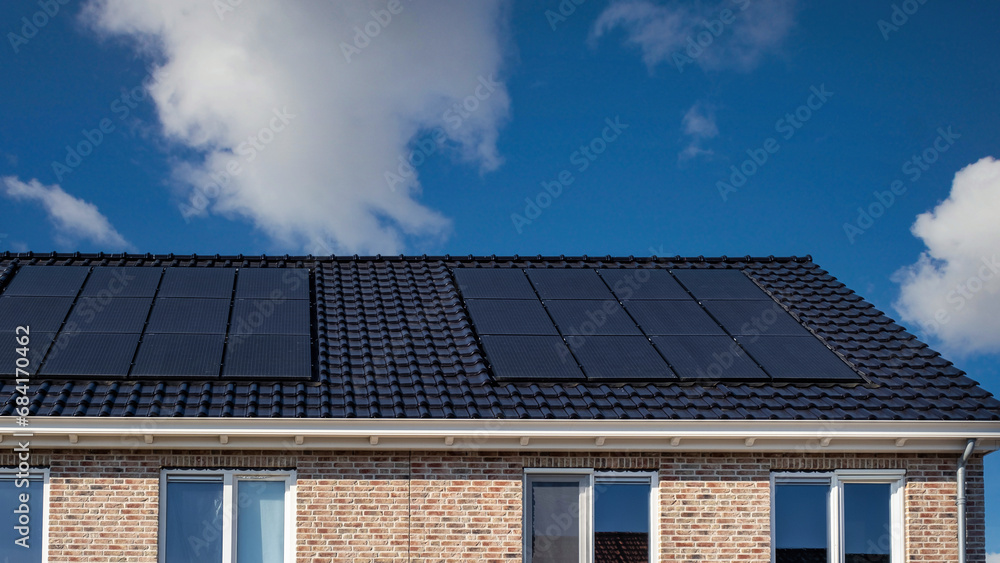 Newly built houses with solar panels attached on the roof against a sunny sky, Close up of new building with black solar panels. Zonnepanelen, Zonne energie, Translation: Solar panel, Sun Energy Newly built houses with solar panels attached on the roof against a sunny sky, Close up of new building with black solar panels. Zonnepanelen, Zonne energie, Translation: Solar panel, Sun Energy
