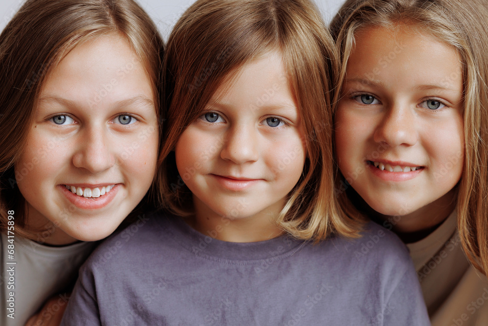 Three blonde sisters in casual clothes smile and hug in front of a white wall. Concept for demonstrating family love, friendship and happiness. Faces in a row. Three blonde sisters in casual clothes smile and hug in front of a white wall. Concept for demonstrating family love, friendship and happiness. Faces in a row.