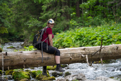 a break in a mountain hike, sitting on a log that has fallen over a stream, a woman looks around