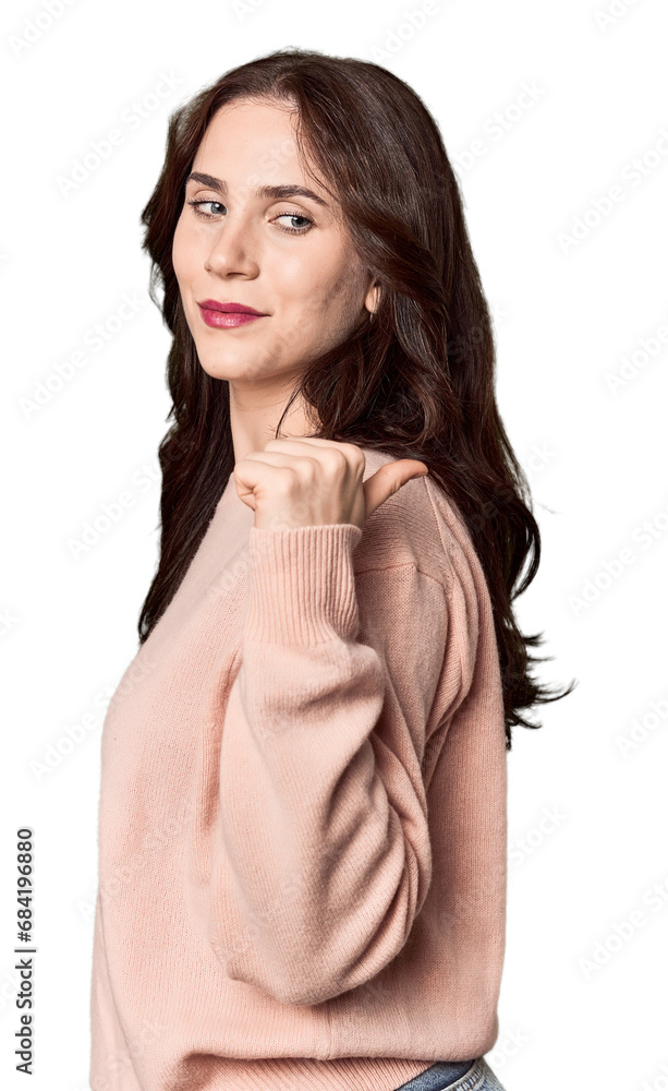 Young Caucasian woman in a studio setting points with thumb finger away, laughing and carefree.