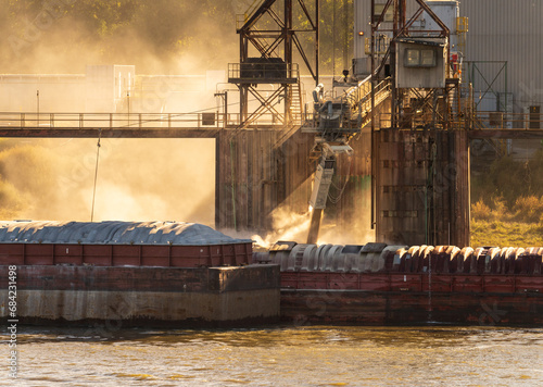 Tugboat pushing freight barges past grain loading dock with dust illuminated by the sun in East St Louis Illinois