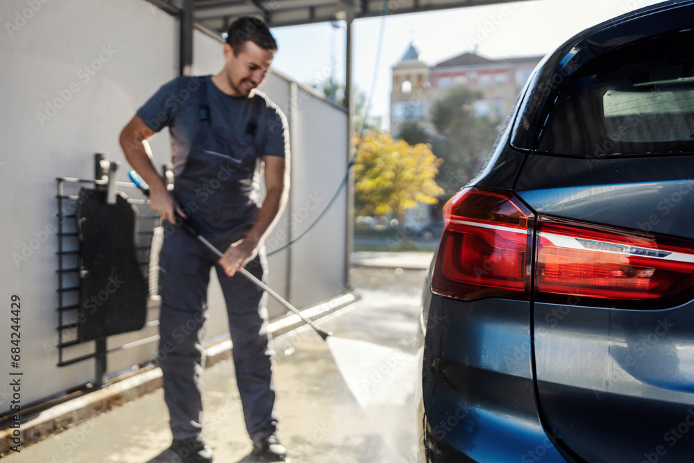 A cheerful car wash station worker cleaning car with water gun.