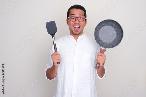 Adult Asian man showing wow expression while holding frying pan and spatula photo