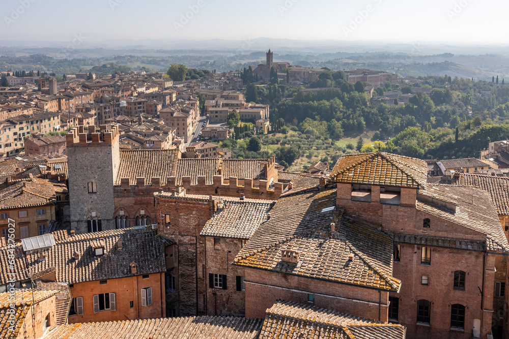 Historical center with tiled roofs, medieval tower and distant hills in morning haze, Siena, Italy