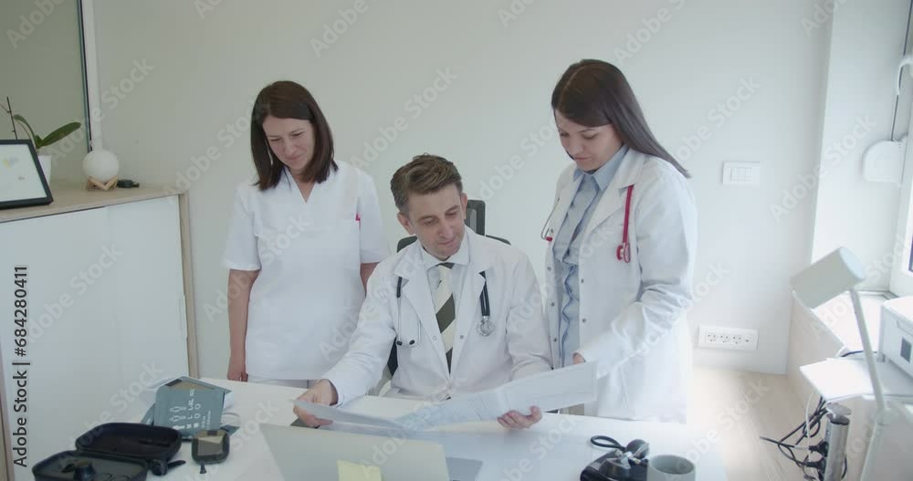 Two medical nurses consult or talk to their experienced male doctor about the results of their patients. Three doctors are analyzing medical results in a small modern office. Two medical nurses consult or talk to their experienced male doctor about the results of their patients. Three doctors are analyzing medical results in a small modern office.