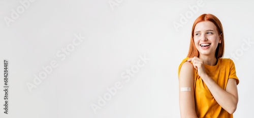 Portrait of a red-haired smiling young woman after getting a vaccine standing on a white background, showing her arm with bandage after receiving vaccination photo