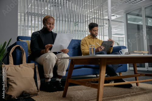 Two multiracial young adults in businesswear reading and using digital tablet on sofa in office