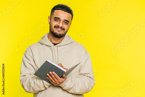 Thoughtful journalist young Indian man making notes, writing down thoughts with pen into notepad notebook diary, to do list, good idea inspiration. Arabian Hindu guy isolated on yellow background