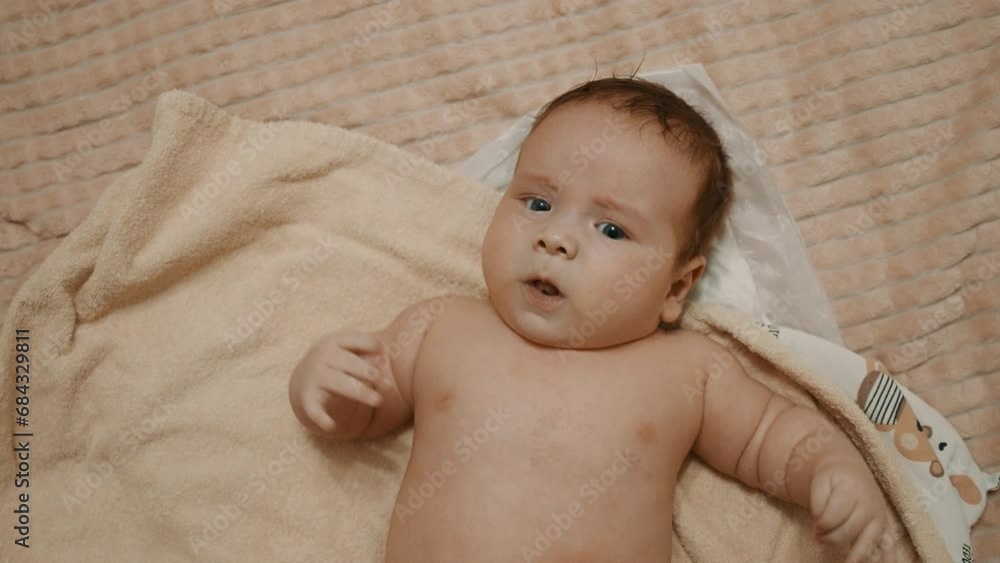 Small baby lies on bed, waves his hands, frowns. Newborn baby lies on a bath baby towel, frowns. Undressed child of European appearance. Negative emotion. Blue eyes. Newborn baby looks with interest Small baby lies on bed, waves his hands, frowns. Newborn baby lies on a bath baby towel, frowns. Undressed child of European appearance. Negative emotion. Blue eyes. Newborn baby looks with interest