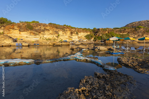 A picturesque bay on the island of Rhodes with shallow water that forms small lagoons between the rocks. There are sunbeds and umbrellas in the water.