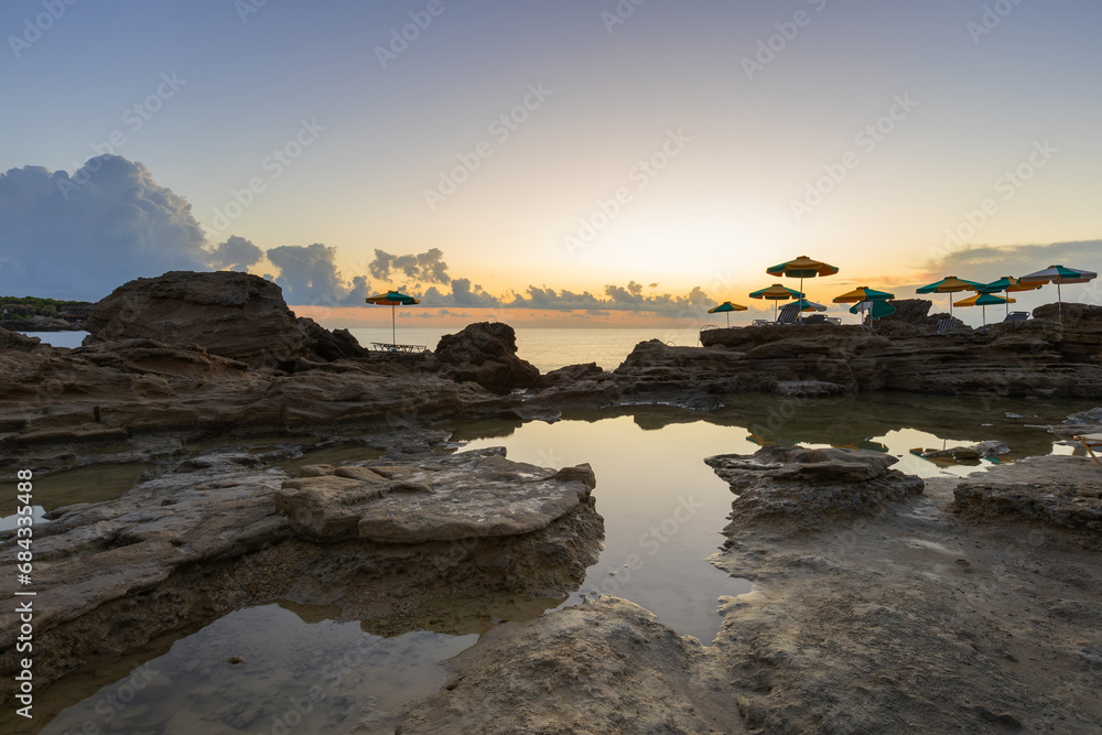 A romantic bay by the sea at sunrise with small lagoons in which the sky is reflected. Among the lagoons, there are umbrellas with sunbeds on the stones.