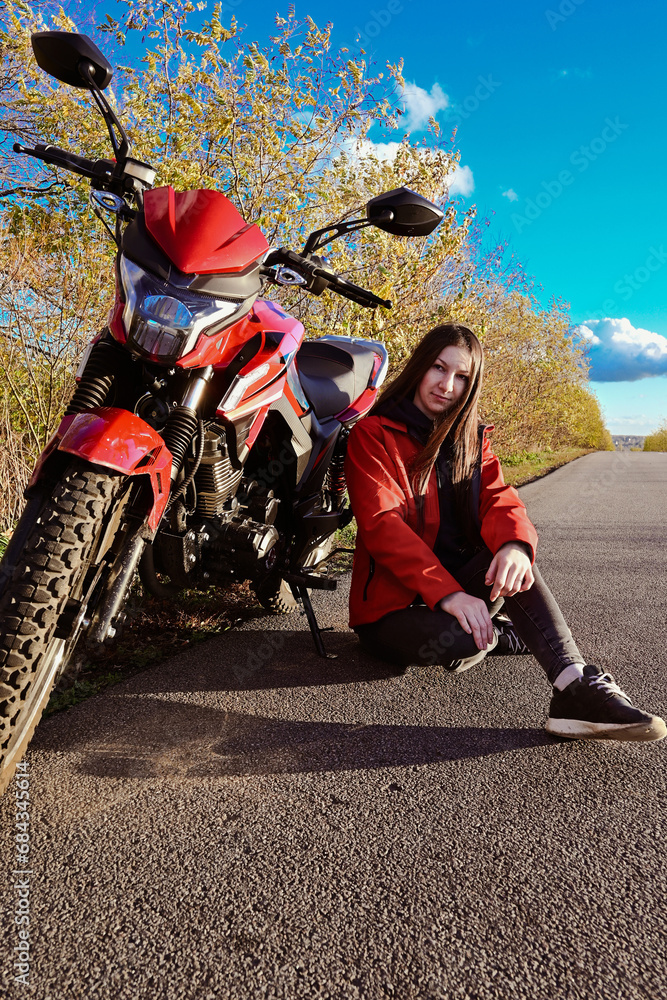 Obraz premium young attractive biker girl in red sitting next to her red motorcycle in the middle of the road. biker with a motorcycle outside the city.